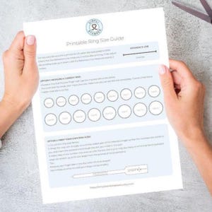 May include: A printable ring size guide held by hands. The guide includes instructions and circles representing ring sizes. The top of the page has the text "Printable Ring Size Guide" and a logo. A reference line measures 2 inches.