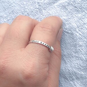 Pu&ograve; includere: Un anello d'argento con la scritta "Just Breathe". L'anello ha un dettaglio avvolto in filo metallico a un'estremit&agrave;. L'anello &egrave; su un dito, su uno sfondo azzurro.