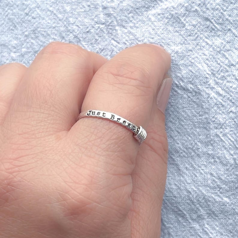 Pu&ograve; includere: Un anello d'argento con la scritta "Just Breathe". L'anello ha un dettaglio avvolto in filo metallico a un'estremit&agrave;. L'anello &egrave; su un dito, su uno sfondo azzurro.