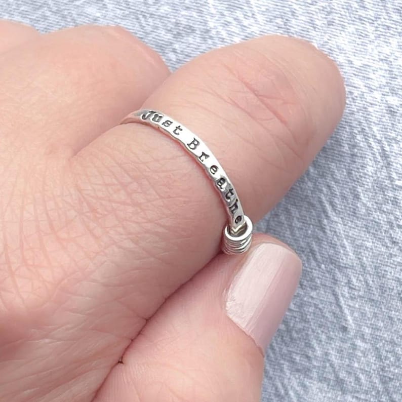 Pu&ograve; includere: Un anello d'argento con le parole "Just Breathe" stampate attorno alla fascia. L'anello &egrave; su un dito e il design include un piccolo dettaglio a spirale nella parte inferiore.