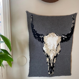 May include: A rectangular crochet wall hanging featuring a large bull skull design. The background is a solid dark gray, while the skull is rendered in shades of cream, white, and black, showcasing the intricate yarn stitches and texture.