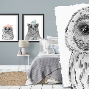 May include: Two framed watercolor prints of woodland animals. One features an owl wearing a pink bow, the other a bear wearing a blue hat. The prints are in black frames and are displayed on a light blue wall.