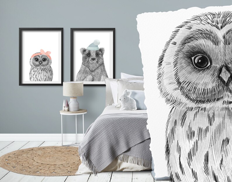 May include: Two framed watercolor prints of woodland animals. One features an owl wearing a pink bow, the other a bear wearing a blue hat. The prints are in black frames and are displayed on a light blue wall.