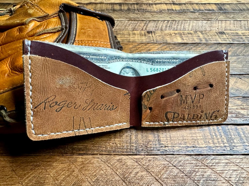 May include: A brown leather wallet with a baseball glove design. The wallet has a brown leather flap with the words "Roger Maris" embossed on it. The inside of the wallet is lined with a burgundy leather. The wallet is open and has a two dollar bill inside. The wallet is sitting on a wooden surface.