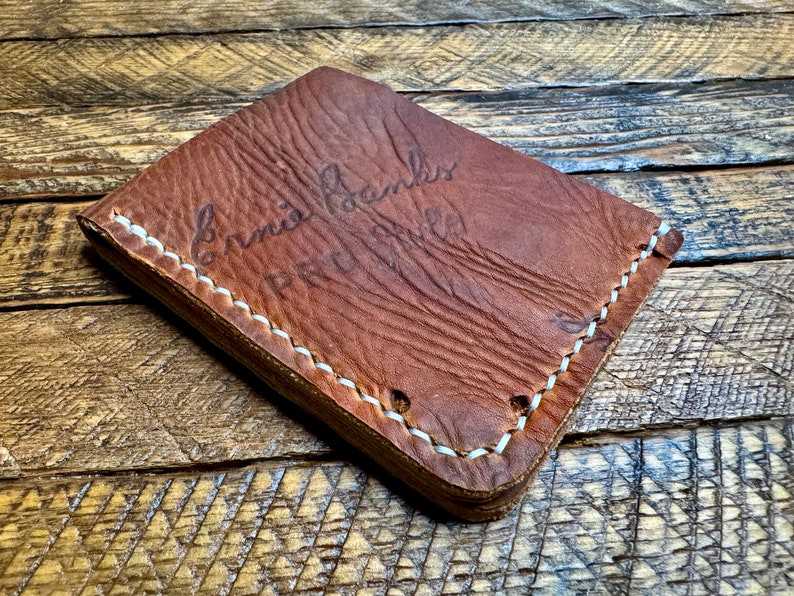 Puede incluir: Una cartera de cuero marr&oacute;n con costuras blancas. La cartera tiene el texto "Ernie Banks" y "Pro Hall" grabado en relieve en la parte delantera.