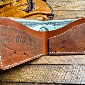 Puede incluir: Una cartera de cuero marr&oacute;n con costuras blancas y el texto "The Big Scoop" grabado en la parte delantera. La cartera est&aacute; abierta y se puede ver un billete de dos d&oacute;lares en el interior.