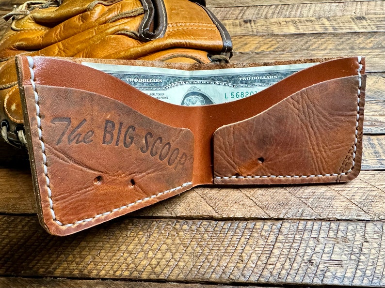 Puede incluir: Una cartera de cuero marr&oacute;n con costuras blancas y el texto "The Big Scoop" grabado en la parte delantera. La cartera est&aacute; abierta y se puede ver un billete de dos d&oacute;lares en el interior.