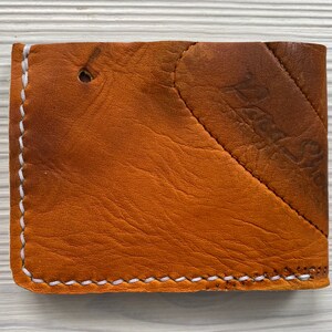 K&ouml;nnte beinhalten: Eine braune Ledergeldb&ouml;rse mit wei&szlig;en N&auml;hten an den Kanten. Die Geldb&ouml;rse hat ein diagonales Design auf der Vorderseite und ein kleines Loch in der N&auml;he der Oberseite. Das Leder hat ein abgenutztes Aussehen.