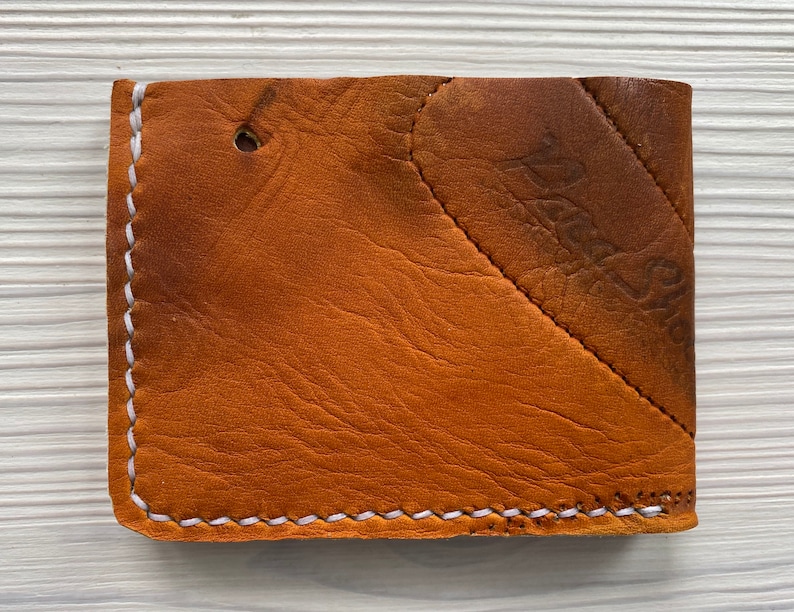 K&ouml;nnte beinhalten: Eine braune Ledergeldb&ouml;rse mit wei&szlig;en N&auml;hten an den Kanten. Die Geldb&ouml;rse hat ein diagonales Design auf der Vorderseite und ein kleines Loch in der N&auml;he der Oberseite. Das Leder hat ein abgenutztes Aussehen.