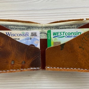 Peut inclure: Un portefeuille en cuir marron avec deux poches, l'une contenant un permis de conduire du Wisconsin et l'autre une carte de d&eacute;bit de Westconsin Credit Union. Le portefeuille a des d&eacute;tails de couture blanche et un aspect us&eacute; et vieilli.