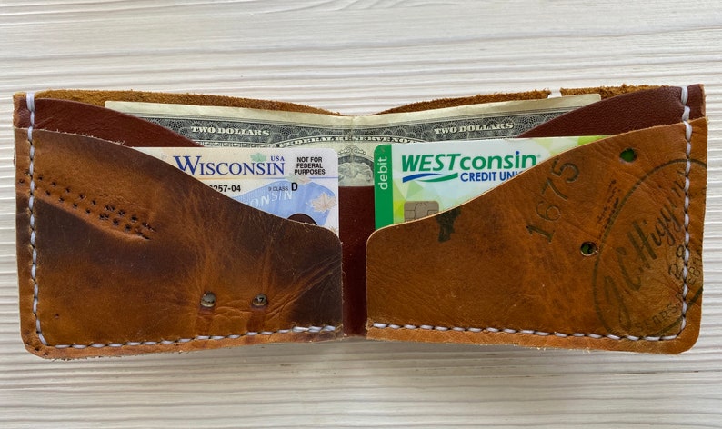 Peut inclure: Un portefeuille en cuir marron avec deux poches, l'une contenant un permis de conduire du Wisconsin et l'autre une carte de d&eacute;bit de Westconsin Credit Union. Le portefeuille a des d&eacute;tails de couture blanche et un aspect us&eacute; et vieilli.