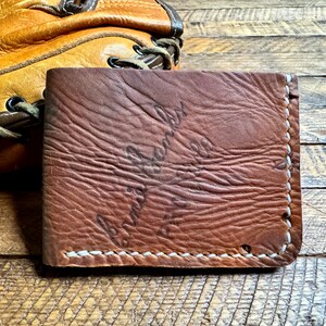 Puede incluir: Una cartera de cuero marr&oacute;n con costuras blancas. La cartera tiene un aspecto desgastado y el texto "Cuero genuino" est&aacute; estampado en la parte delantera.