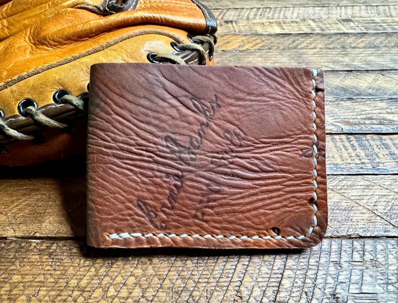 Puede incluir: Una cartera de cuero marr&oacute;n con costuras blancas. La cartera tiene un aspecto desgastado y el texto "Cuero genuino" est&aacute; estampado en la parte delantera.