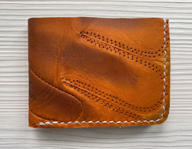 K&ouml;nnte beinhalten: Ein braunes Lederportemonnaie mit zweifachem Design. Das Portemonnaie hat dekorative N&auml;hte und einen wei&szlig; gen&auml;hten Rand. Das Leder hat ein abgenutztes Aussehen, wobei dunklere Bereiche dem Design Tiefe verleihen. Ein klassisches Accessoire.