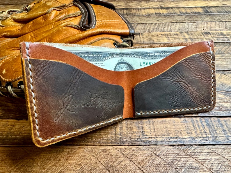 May include: A brown leather wallet with a dark brown leather interior. The wallet is open and has a two pound note inside. The wallet has white stitching around the edges and a signature on the front.