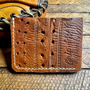 Puede incluir: Una cartera de cuero marr&oacute;n con costuras blancas y un aspecto desgastado. La cartera tiene un dise&ntilde;o &uacute;nico con agujeros perforados en todo el cuero. El texto "Grip It" est&aacute; estampado en el lado derecho de la cartera.