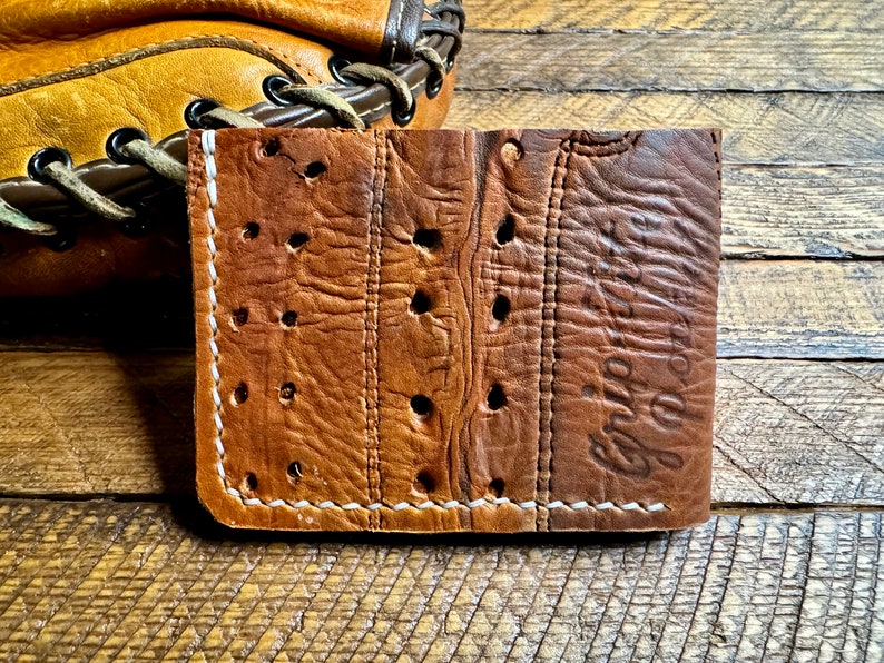 Puede incluir: Una cartera de cuero marr&oacute;n con costuras blancas y un aspecto desgastado. La cartera tiene un dise&ntilde;o &uacute;nico con agujeros perforados en todo el cuero. El texto "Grip It" est&aacute; estampado en el lado derecho de la cartera.