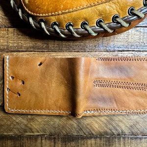 Puede incluir: Una cartera de cuero marr&oacute;n con costuras blancas. La cartera est&aacute; doblada por la mitad y est&aacute; sobre una superficie de madera.