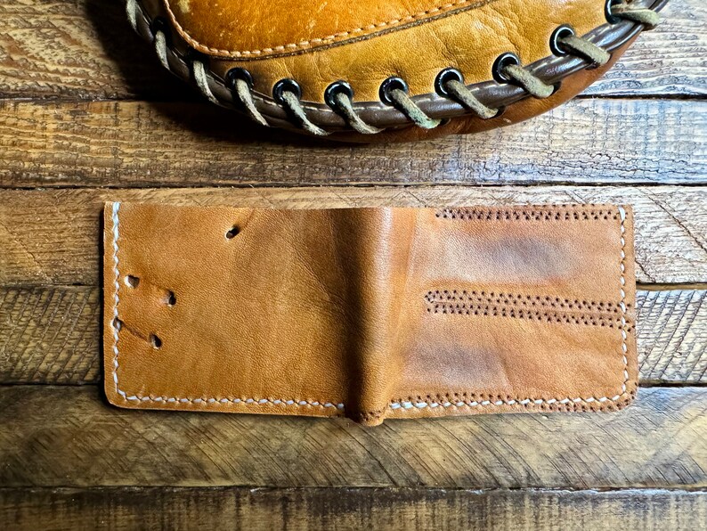Puede incluir: Una cartera de cuero marr&oacute;n con costuras blancas. La cartera est&aacute; doblada por la mitad y est&aacute; sobre una superficie de madera.