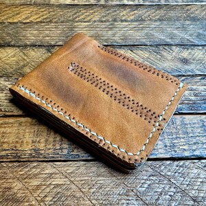 Puede incluir: Una cartera de cuero marr&oacute;n con costuras blancas. La cartera est&aacute; doblada y apoyada sobre una superficie de madera.