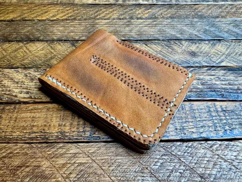 Puede incluir: Una cartera de cuero marr&oacute;n con costuras blancas. La cartera est&aacute; doblada y apoyada sobre una superficie de madera.