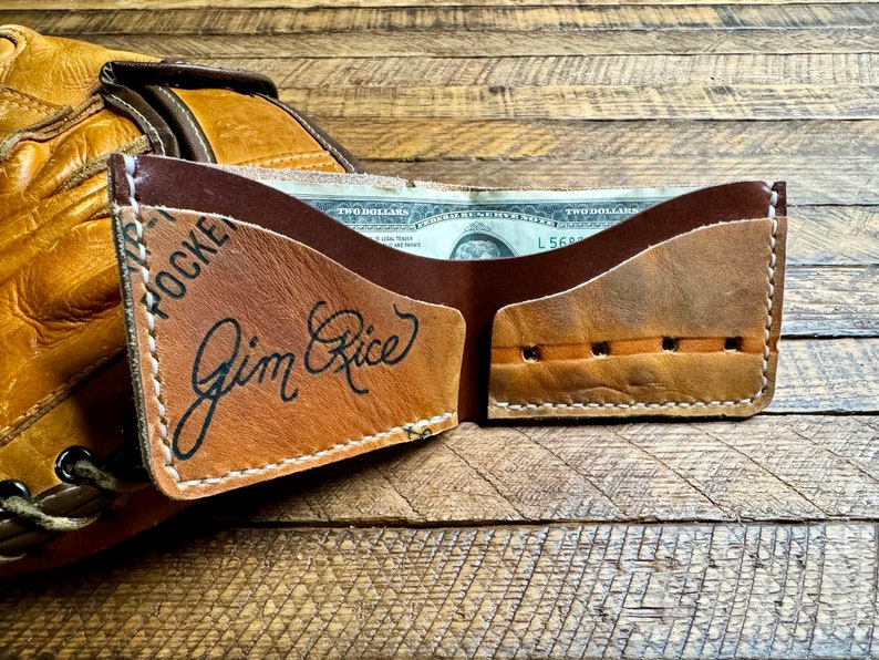 Puede incluir: Una cartera de cuero marr&oacute;n con una solapa de cuero tostado. La solapa tiene el texto "Jim Rice" escrito en tinta negra. La cartera est&aacute; abierta y se puede ver un billete de dos d&oacute;lares en el interior.