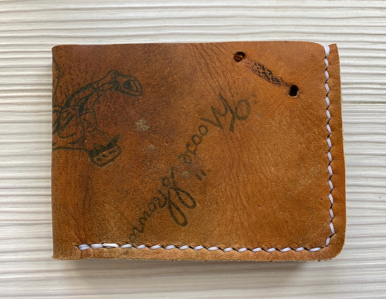 Peut inclure: Un portefeuille en cuir marron avec des coutures blanches et un graphique noir d'une main tenant une cl&eacute;. Le portefeuille porte le texte "always do you".