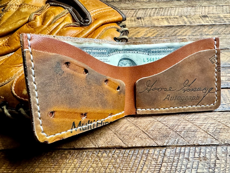 May include: A brown leather wallet with a stitched edge and a white inscription that reads "Multi Hip". The wallet is open and has a two dollar bill inside. The wallet has a brown leather flap with a white inscription that reads "Loose Change Autograph".