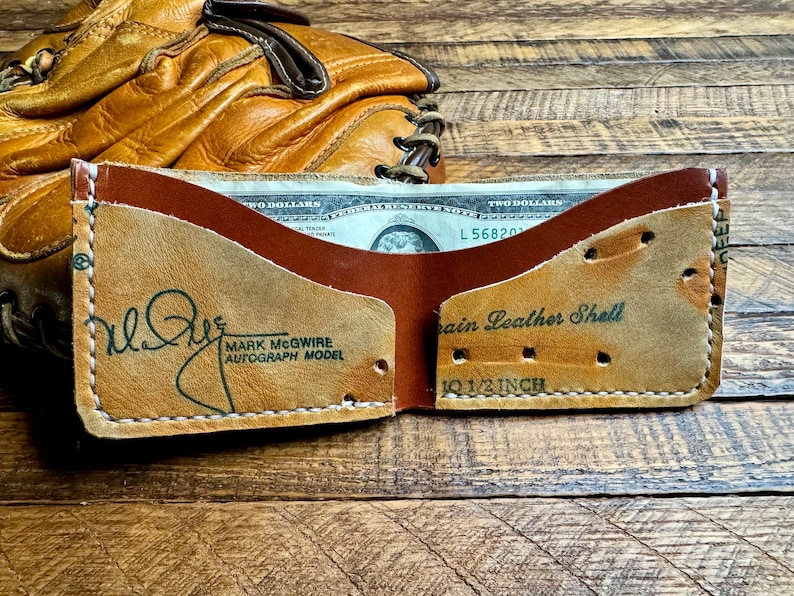 Pu&ograve; includere: Un portafoglio a due ante in pelle di guanto da baseball, con interno ed esterno marrone. Il portafoglio presenta la scritta "MARK MCGWIRE AUTOGRAPH MODEL" e "Grain Leather Shell". Il portafoglio &egrave; lungo 26,67 cm.