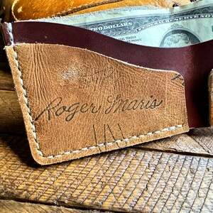 May include: A brown leather wallet with a white stitch detail. The wallet has a burgundy leather flap and the name "Roger Maris" is written in black ink on the front.