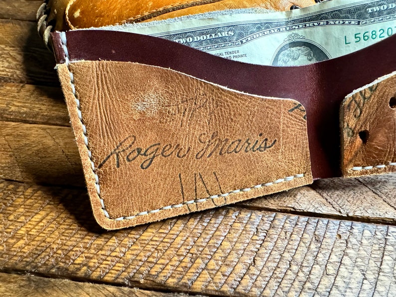 May include: A brown leather wallet with a white stitch detail. The wallet has a burgundy leather flap and the name "Roger Maris" is written in black ink on the front.