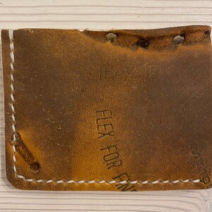 Puede incluir: Una cartera de cuero marr&oacute;n con costuras blancas y el texto "Flex For Fin" en relieve en la parte delantera.