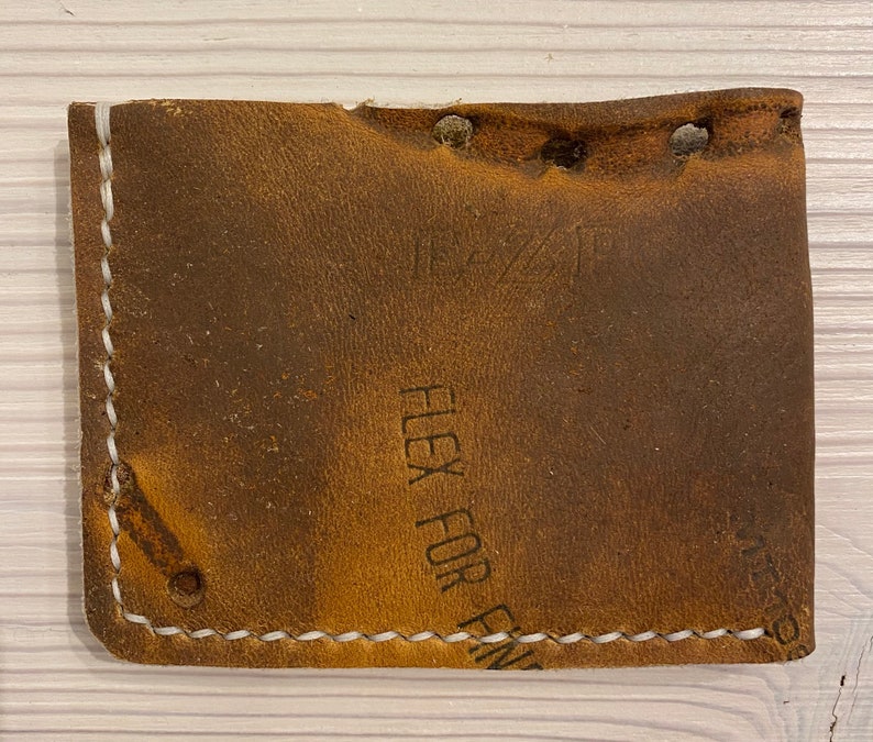 Puede incluir: Una cartera de cuero marr&oacute;n con costuras blancas y el texto "Flex For Fin" en relieve en la parte delantera.