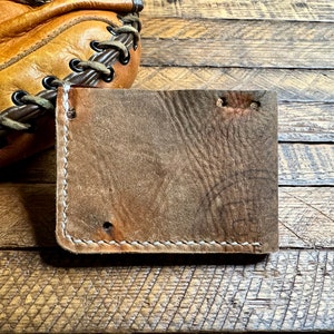 Puede incluir: Una cartera de cuero marr&oacute;n con costuras blancas. La cartera est&aacute; sobre una superficie de madera.