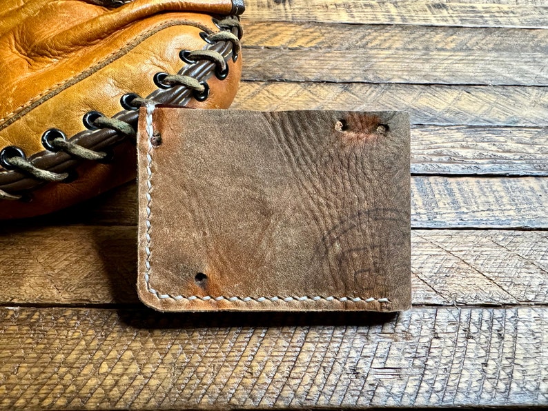 Puede incluir: Una cartera de cuero marr&oacute;n con costuras blancas. La cartera est&aacute; sobre una superficie de madera.