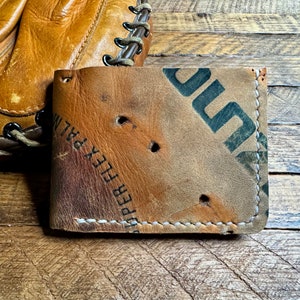 Pu&ograve; includere: Portafoglio a due ante in pelle beige, realizzato con un guanto da baseball riciclato. Il portafoglio presenta cuciture visibili e la scritta "SUPER FLEX PALM". Il design ha un aspetto invecchiato. Un guanto da baseball &egrave; visibile sullo sfondo.