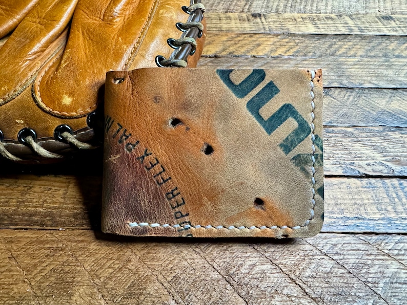 Pu&ograve; includere: Portafoglio a due ante in pelle beige, realizzato con un guanto da baseball riciclato. Il portafoglio presenta cuciture visibili e la scritta "SUPER FLEX PALM". Il design ha un aspetto invecchiato. Un guanto da baseball &egrave; visibile sullo sfondo.