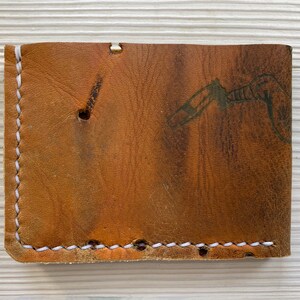 Peut inclure: Un portefeuille en cuir marron avec des coutures blanches. Le portefeuille a un aspect us&eacute; et un motif discret sur le devant.