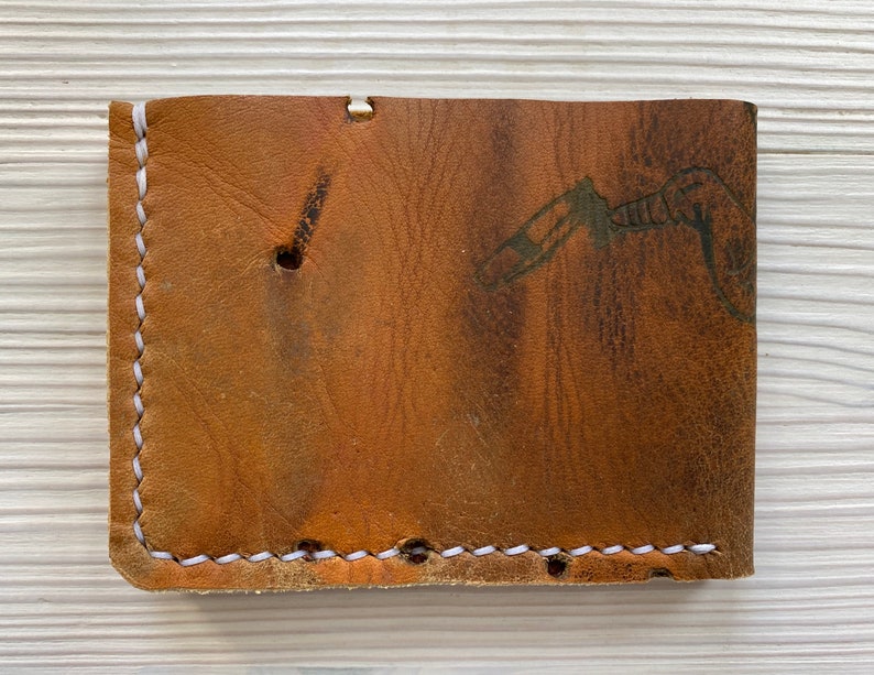 Peut inclure: Un portefeuille en cuir marron avec des coutures blanches. Le portefeuille a un aspect us&eacute; et un motif discret sur le devant.