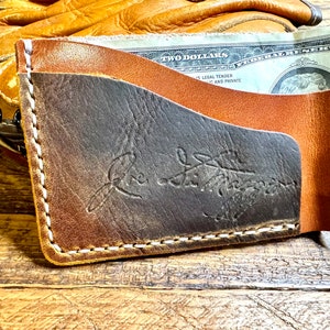 May include: A brown leather wallet with a stitched edge and a black leather interior. The wallet has a logo embossed on the front that reads "The Leather Works".