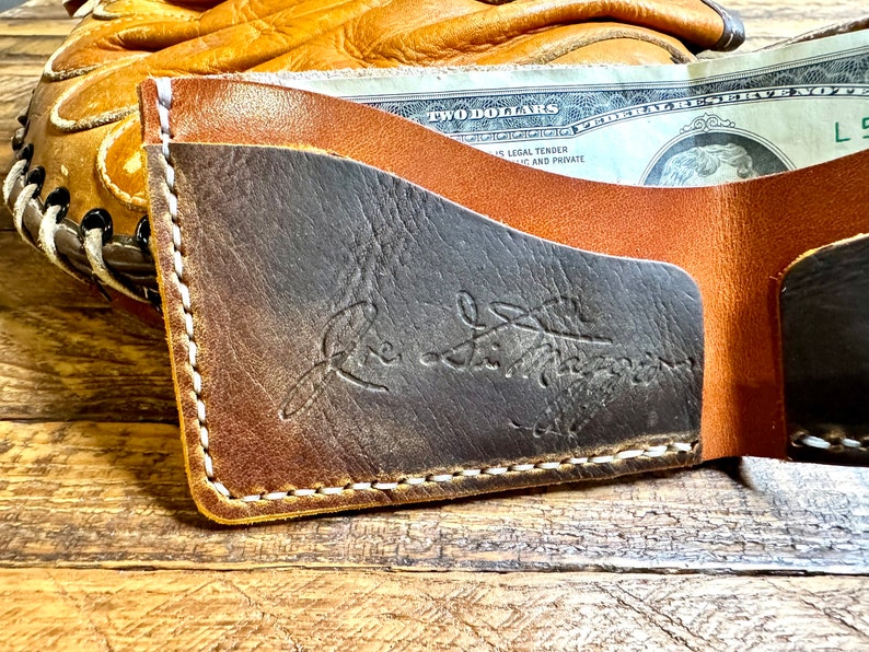 May include: A brown leather wallet with a stitched edge and a black leather interior. The wallet has a logo embossed on the front that reads "The Leather Works".