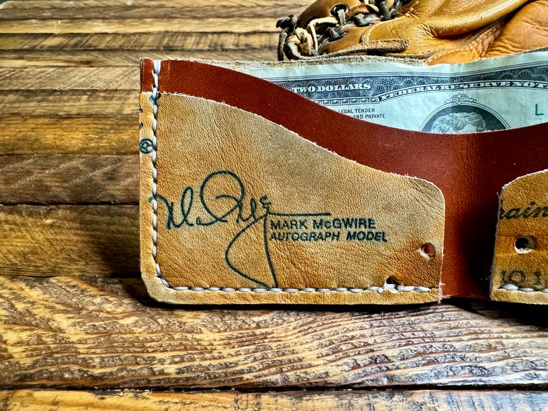 Pu&ograve; includere: Un portafoglio in pelle marrone con un guanto da baseball sullo sfondo. Il portafoglio ha un esterno marrone chiaro con l'autografo di Mark McGwire e la scritta "AUTOGRAPH MODEL". All'interno &egrave; visibile una banconota da due dollari.