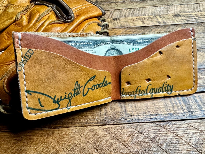 Puede incluir: Una cartera de cuero marr&oacute;n con una inscripci&oacute;n verde que dice "Dwight Gooden" y "Calidad artesanal". La cartera est&aacute; abierta y se puede ver un billete de dos d&oacute;lares en el interior.