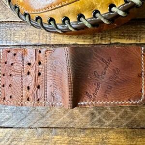 Puede incluir: Una cartera de cuero marr&oacute;n con una costura blanca alrededor de los bordes. La cartera est&aacute; doblada por la mitad y el interior es visible. El interior de la cartera tambi&eacute;n es de cuero marr&oacute;n y tiene las palabras "Ernie Banks" y "Pro Style" escritas con tinta negra.