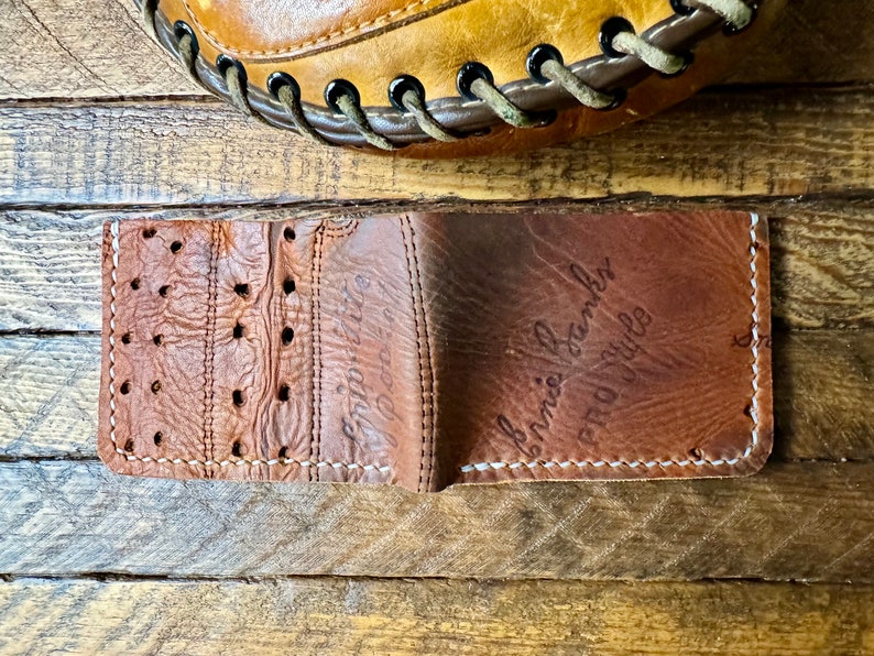 Puede incluir: Una cartera de cuero marr&oacute;n con una costura blanca alrededor de los bordes. La cartera est&aacute; doblada por la mitad y el interior es visible. El interior de la cartera tambi&eacute;n es de cuero marr&oacute;n y tiene las palabras "Ernie Banks" y "Pro Style" escritas con tinta negra.