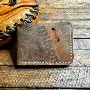 Puede incluir: Una cartera de cuero marr&oacute;n con costuras blancas y el texto "Wilson" estampado en la parte delantera. La cartera est&aacute; sobre una superficie de madera.