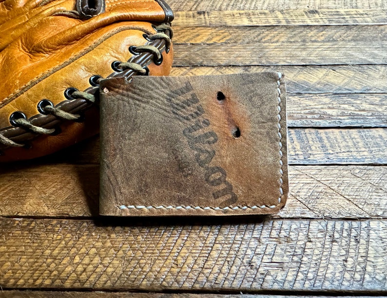 Puede incluir: Una cartera de cuero marr&oacute;n con costuras blancas y el texto "Wilson" estampado en la parte delantera. La cartera est&aacute; sobre una superficie de madera.