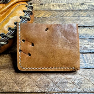 Puede incluir: Una cartera de cuero marr&oacute;n con costuras blancas y un aspecto desgastado. La cartera tiene varios agujeros perforados en el cuero.