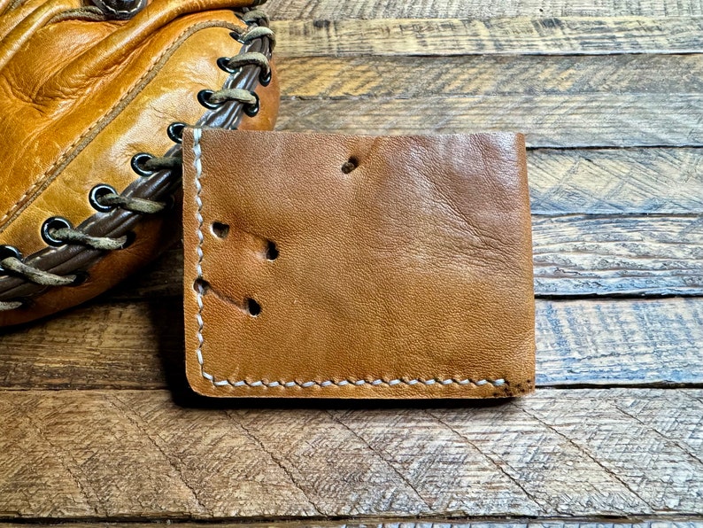 Puede incluir: Una cartera de cuero marr&oacute;n con costuras blancas y un aspecto desgastado. La cartera tiene varios agujeros perforados en el cuero.