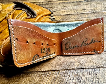 Peut inclure: Un portefeuille biface en cuir marron avec des coutures blanches, avec le texte "1205 Rawlings" et "Dave Parker". Le portefeuille est ouvert, révélant un billet de deux dollars. Un gant de baseball est en arrière-plan.